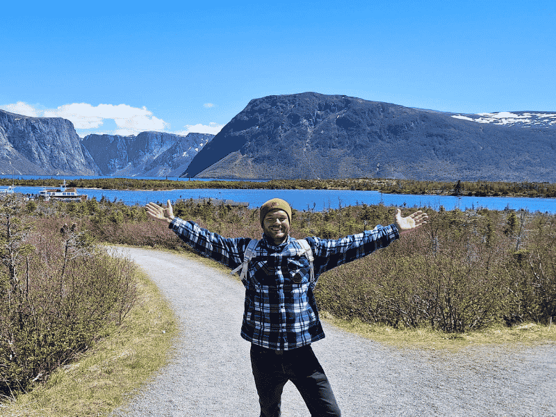Reisespezialist Yannick steht mit weit ausgebreiteten Armen vor einem See und Bergen