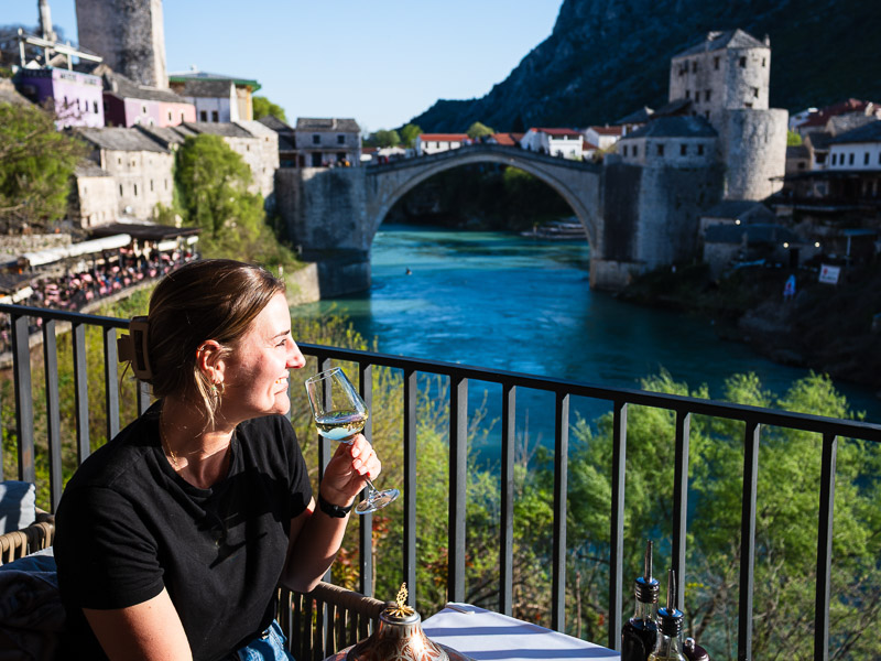Frau sitzt mit einem Glass Wein am Fluss mit Blick auf die alte Brücke in Mostar