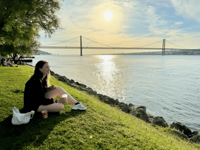 Frau sitzt am Ufer des Flusses Tejo mit der Lissabons Hängebrücke im Hintergrund