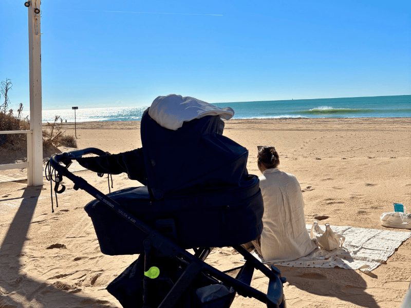 Schwarzer Kinderwagen auf dem Strand, dahinter eine Frau auf einer Decke von hinten, in der Ferne das Meer und blauer Himmel