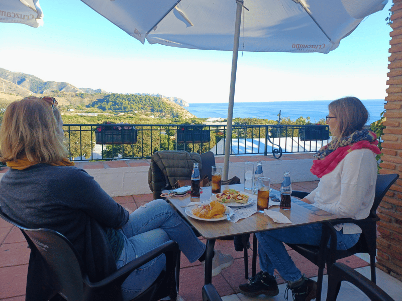 Zwei Frauen am Tisch in einem Restaurant blicken von der Terrasse auf die Berge und das Meer