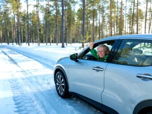 Frau winkt aus Fenster eines silbernen Autos, das über Schnee entlang von Bäumen fährt