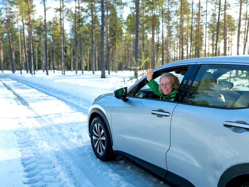 Frau winkt aus Fenster eines silbernen Autos, das über Schnee entlang von Bäumen fährt