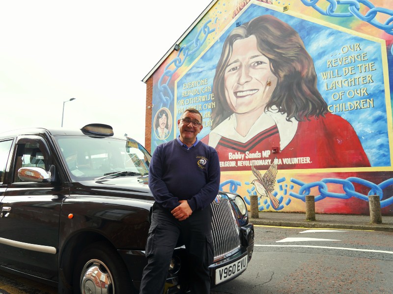 Black cab Guide vor seinem Fahrzeug mit Wandmalerei im Hintergrund.