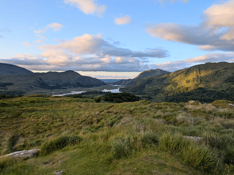 Grün bewachsene Hügel und kleine Berge mit einem Fluss und Wolken am Himmel