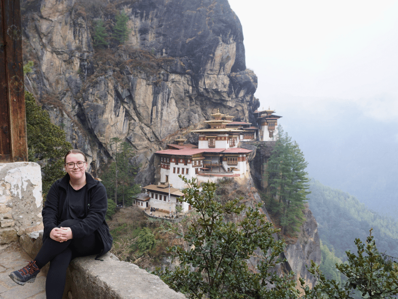 Schwarz gekleidete Frau mit Brille sitzt auf Mauer mit Tigernest in Bhutan im Hintergrund