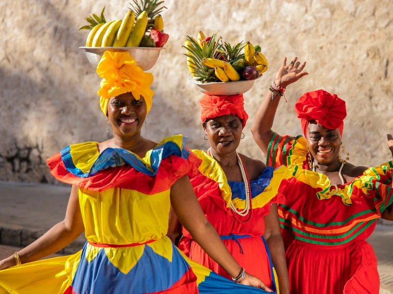 Drei Frauen mit bunten traditionellen Kleidern tragen Schalen voller Früchte auf ihren Köpfen.