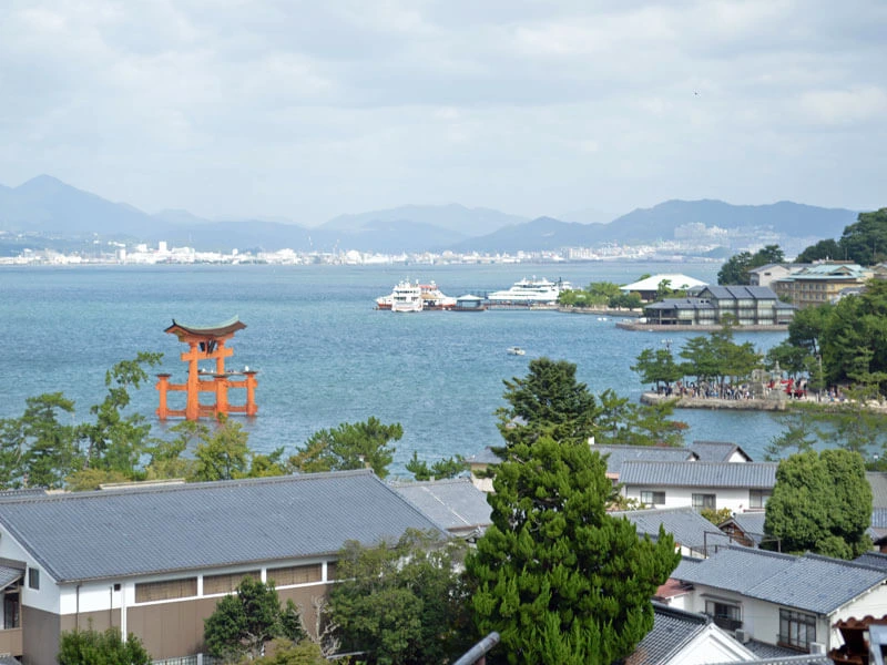Blick vom Hügel auf Miyajima auf das große rote Torii im Wasser mit Hiroshima im Hintergrund