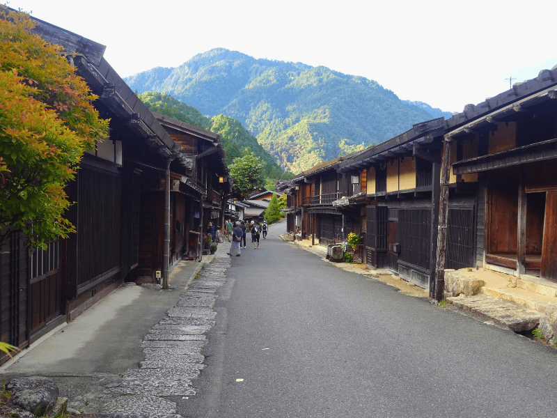 Straße durch ein japanisches Dorf, mit Häusern am Rand der Straße, Menschen und Berge im Hintergrund