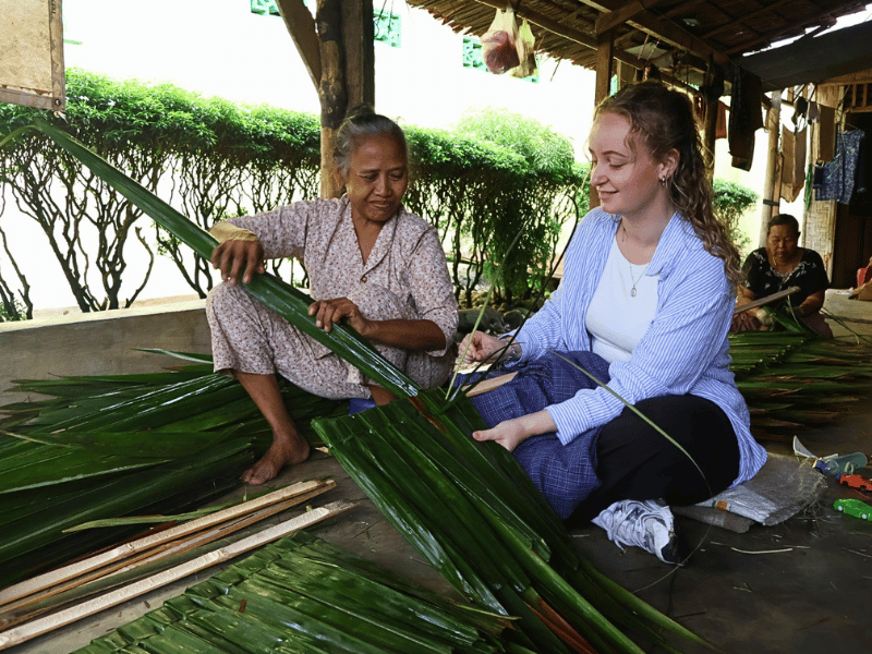 Touristin hilft einheimischer Frau beim Herstellen eines Palmdachs bei Bukit Lawang, Indonesien