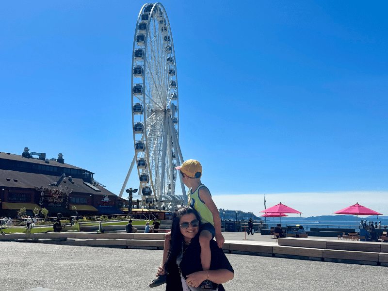 Frau mit Kind auf den Schultern mit Riesenrad im Hintergrund am Hafen in Seattle