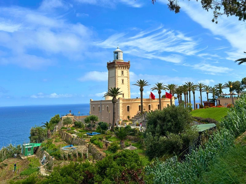 Leuchtturm auf einer Klippe an der Küste von Tanger in Marokko mit Blick auf das Meer, umgeben von Palmen, Gärten und einem strahlend blauen Himmel mit weißen Wolken.