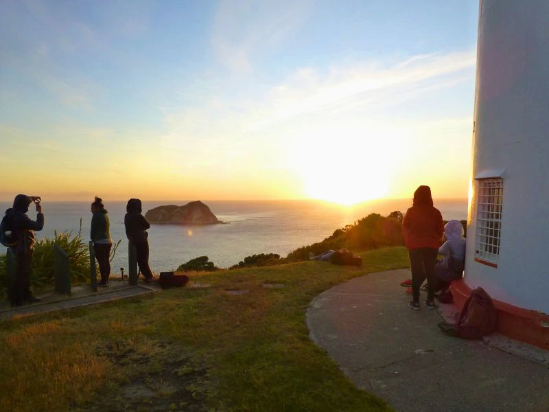 Personen stehen am Leuchtturm und betrachten den Sonnenaufgang am East Cape in Neuseeland.