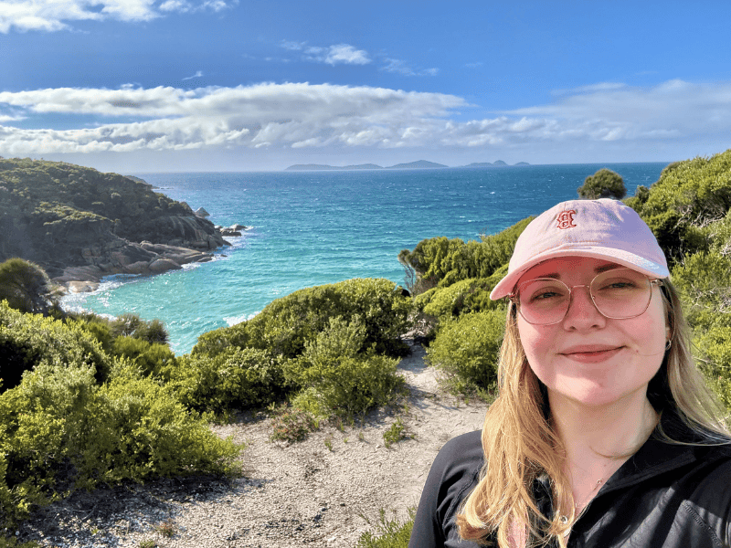 Frau mit Brille vor grünen Büschen, dahinter das Meer und in weiter Ferne Berge