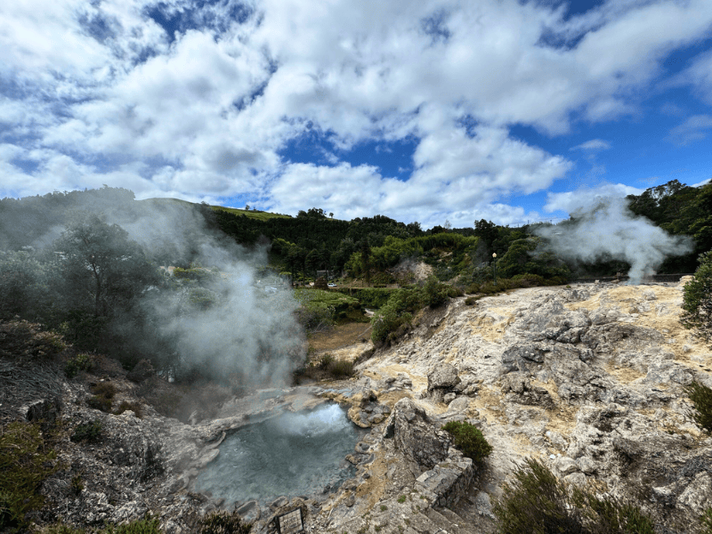 Heiße Quelle mit Dampf inmitten grüner Hügel mit blauem Himmel
