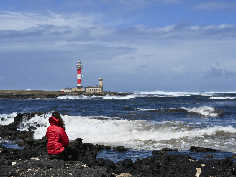 Frau in roter Jacke sitzt auf Steinen am Meer, mit Blick auf rot-weiß gestreiften Leuchtturm
