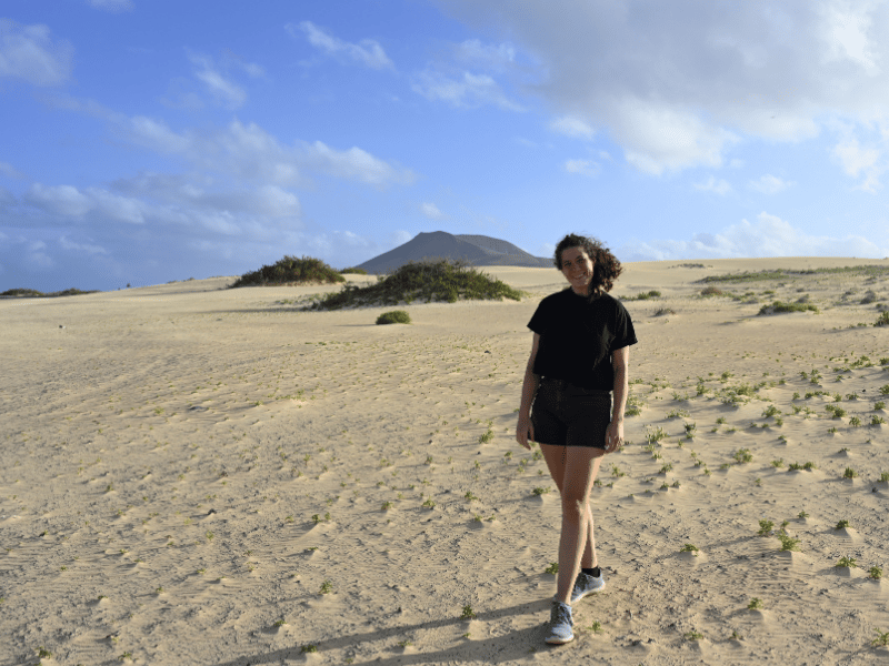 Frau läuft über Sanddünen mit grünen Büscheln und etwas größerer grüner Vegetation im Hintergrund