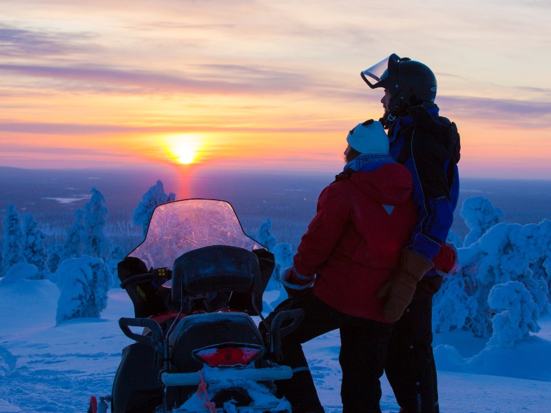 Reisende mit Schneescooter beim Sonnenuntergang, Finnland